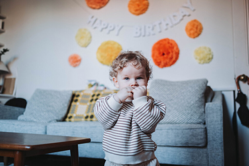 Little boy wearing a striped jumper stands in front of paper decorations that say "Happy Birthday!" 