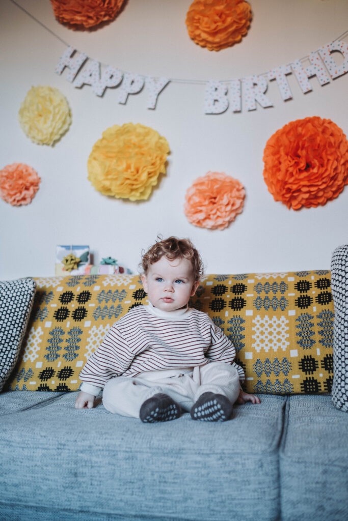 A curly haired toddler wearing a striped shirt sits on his couch with birthday decorations around him 