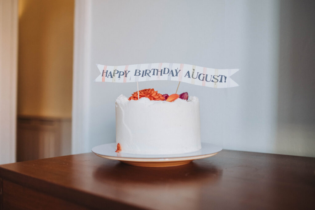 A birthday cake sits on a wooden table, with a decoration that says "Happy Birthday August!" 