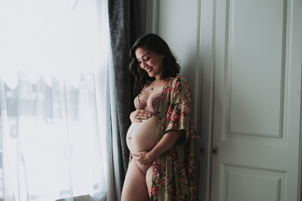 Brown haired woman wearing a floral robe holds her pregnant belly in her bedroom at home in London